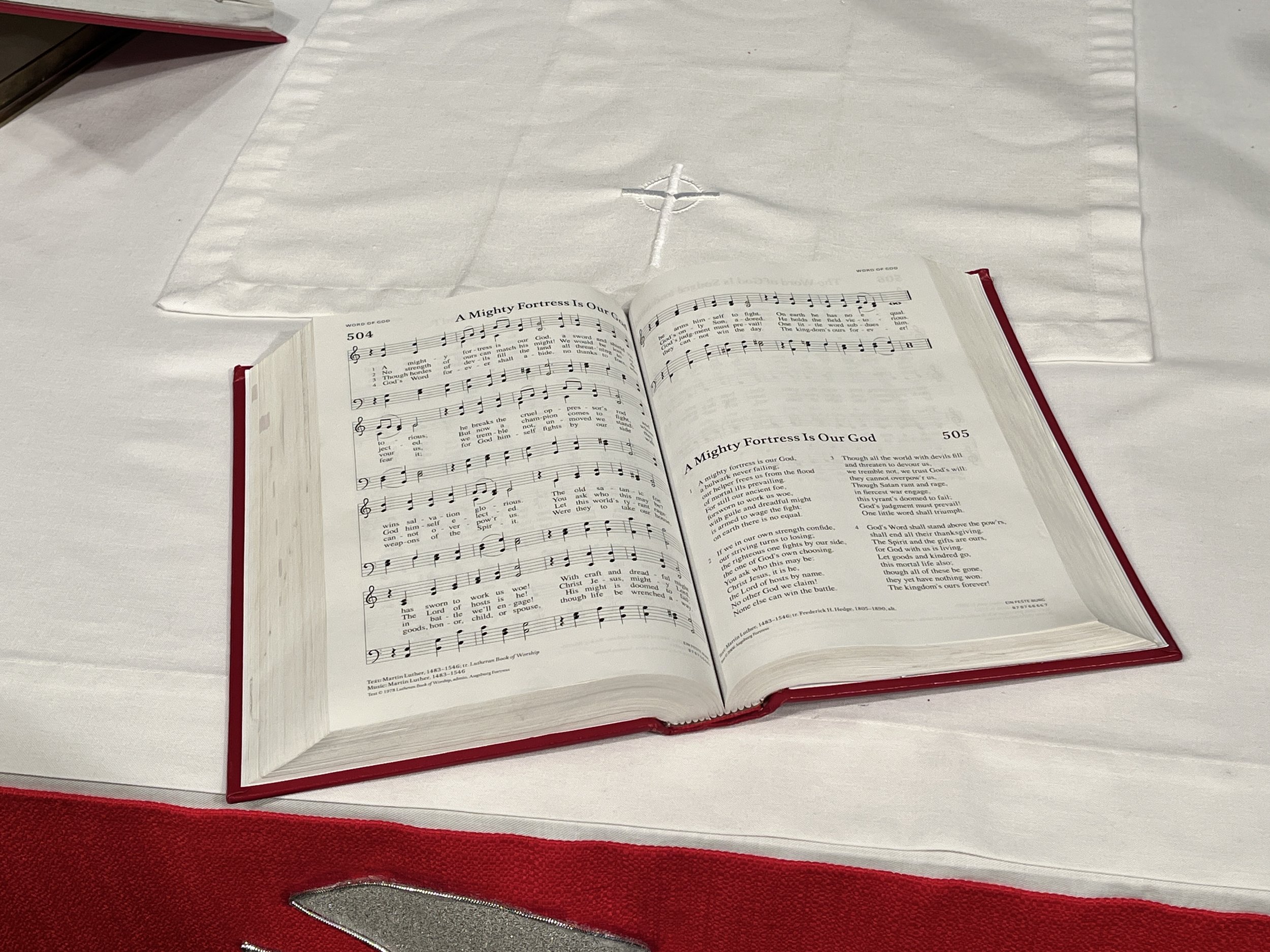 An open hymnal book on a church altar with a white cloth, a cross in the background, and a red border on the altar cloth.