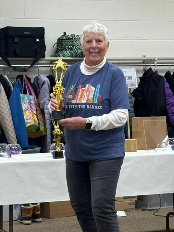 A woman with short white hair, wearing a blue T-shirt and white long-sleeve shirt, holding a trophy, standing in front of a table with awards and clothing in the background.