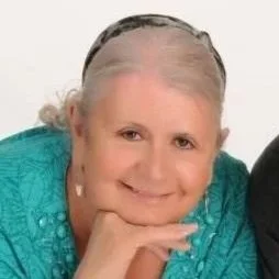 Close-up of an elderly woman with gray hair smiling, resting her chin on her hand, wearing a turquoise top.