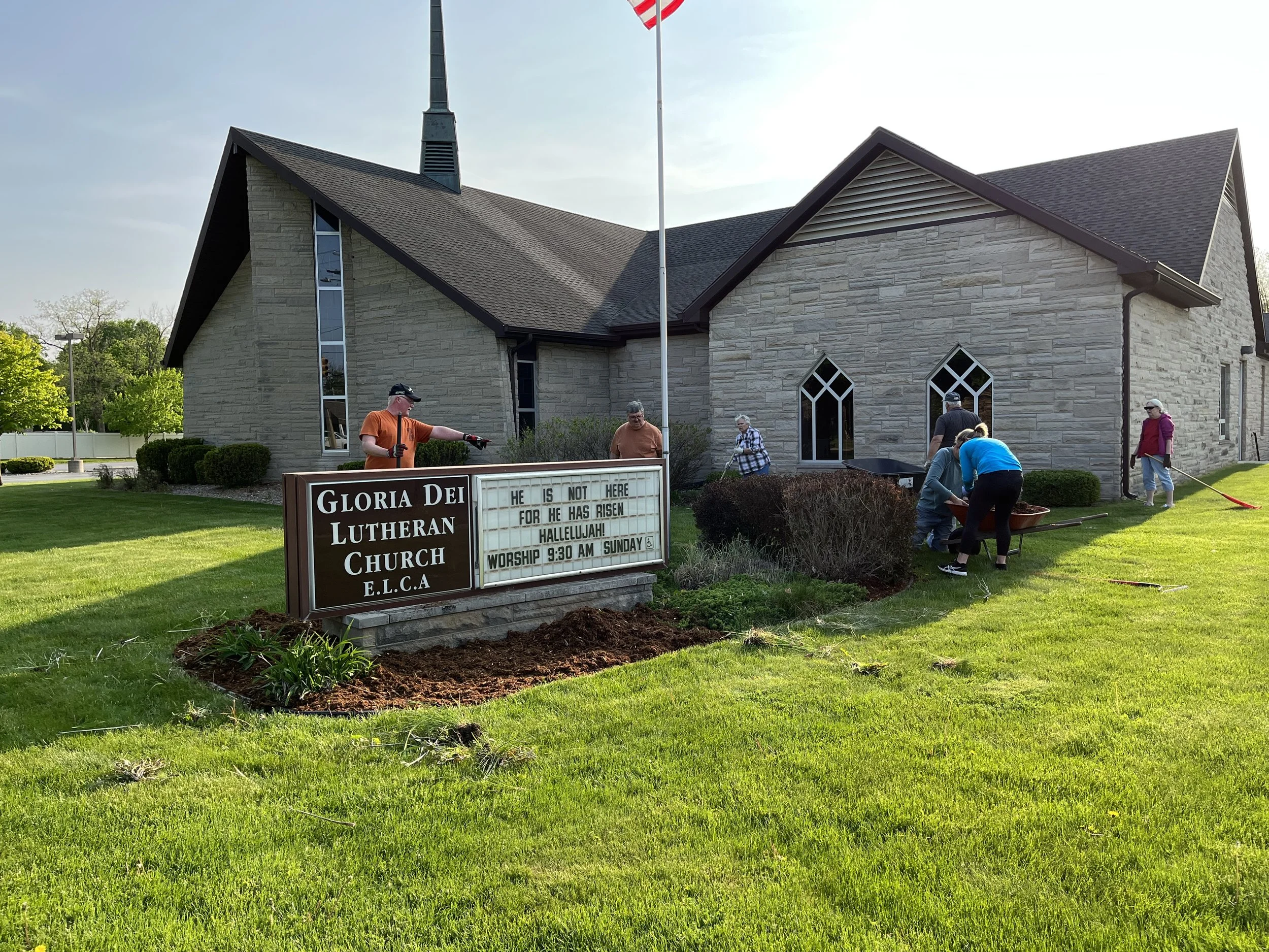People planting flowers outside Gloria Dei Lutheran Church, a stone building with pointed windows, during daylight. A sign displays service information.
