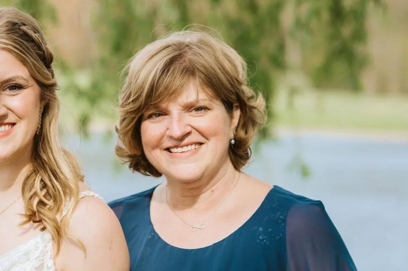 A smiling woman with short blonde hair wearing a dark blue dress, standing outdoors near a body of water with trees in the background.