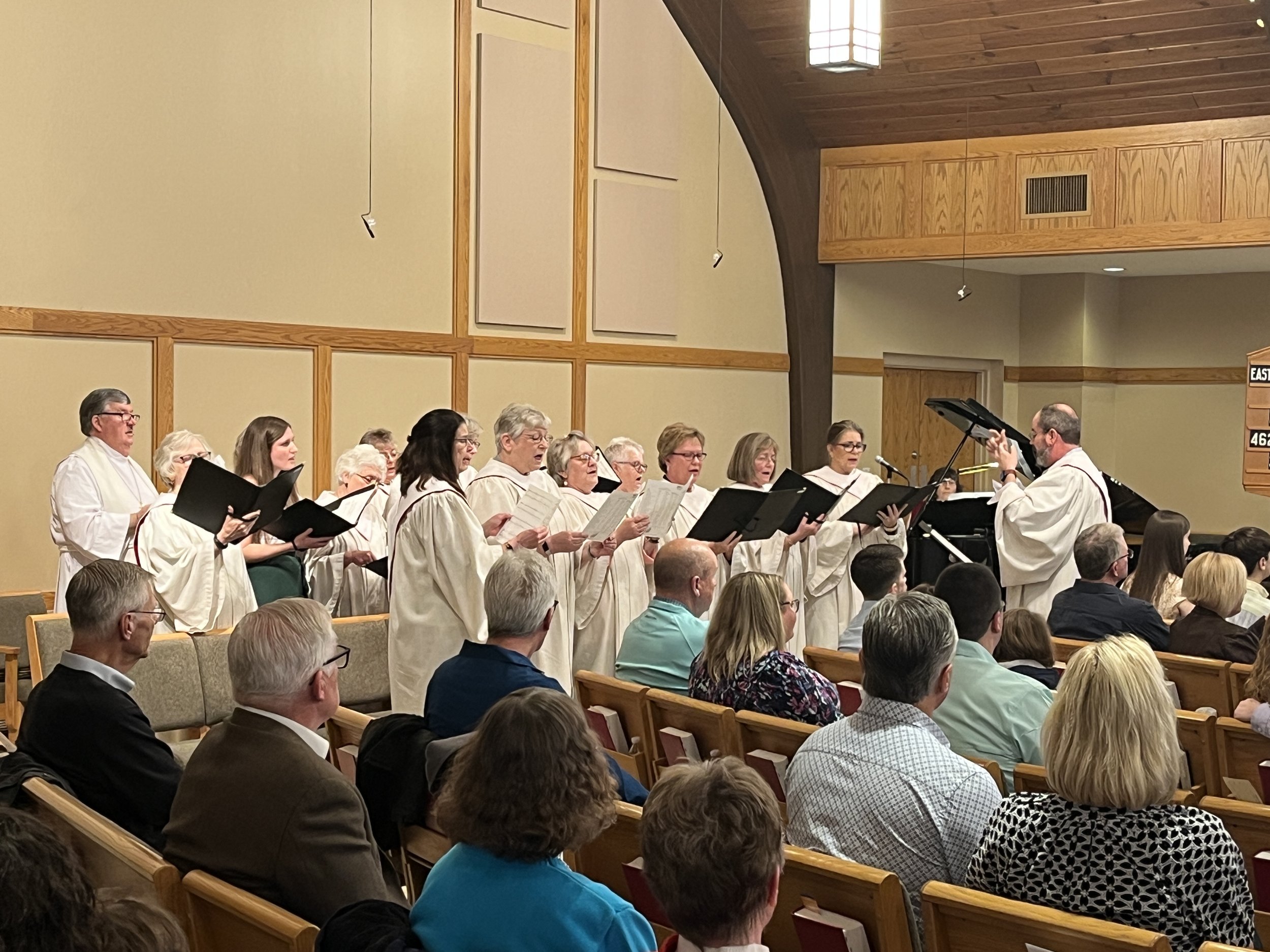 Choir group singing in a church or auditorium, directed by a conductor, with audience members seated in the foreground.