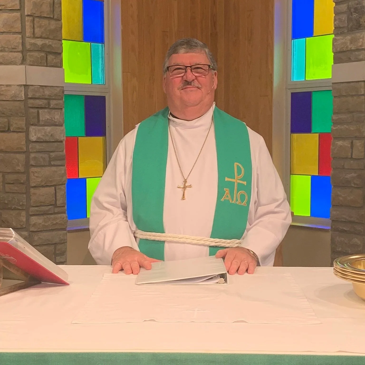 A man dressed as a priest stands at an altar in a church, wearing a white robe with a green stole adorned with religious symbols, a gold cross necklace, and a white rope belt. Behind him are colorful stained glass windows.