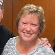 Smiling elderly woman with short blonde hair, wearing a patterned top, in a wooden-paneled room.