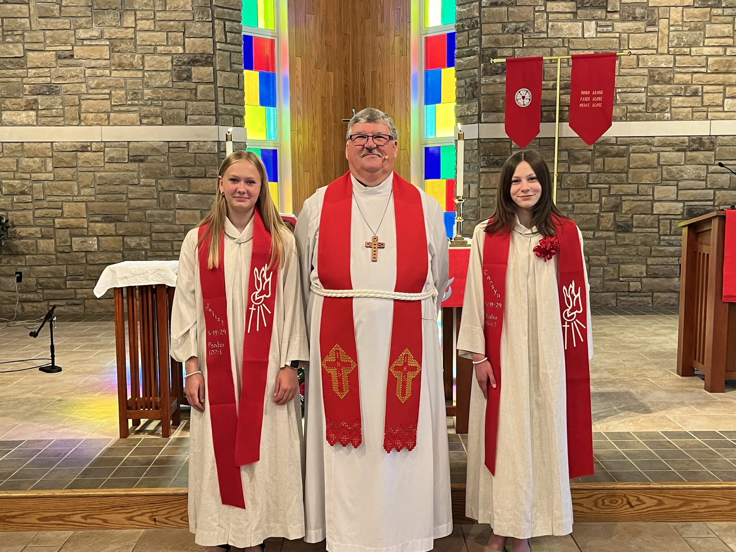 A priest and two young women standing in a church altar, all dressed in white robes with red stoles, with a stained glass window behind them.
