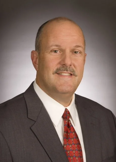 Portrait of a middle-aged man in a gray suit, white shirt, and red patterned tie, with a mustache and short hair, against a gray gradient background.