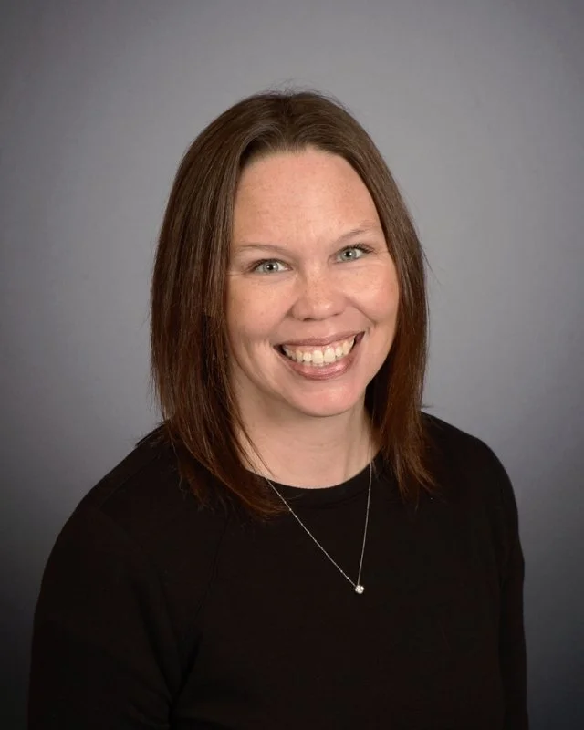 Portrait of a woman with shoulder-length brown hair wearing a black top and a silver necklace, smiling against a gray background.