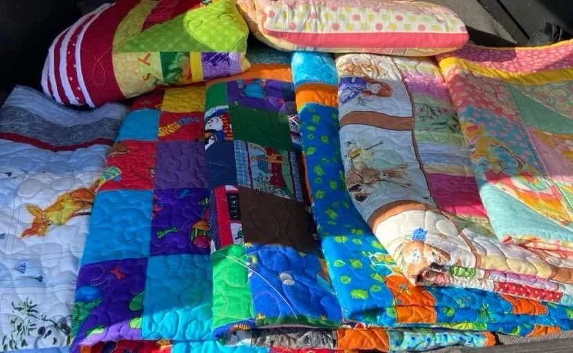 A collection of colorful quilts and blankets, some with children's characters and patterns, folded and stacked on a surface.