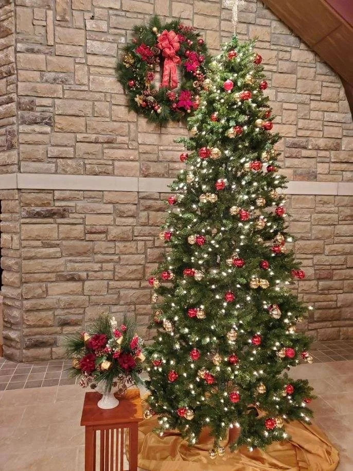 A decorated Christmas tree with ornaments and lights, a wreath with a red bow and flowers hanging on a brick wall, and a flower arrangement on a small table.