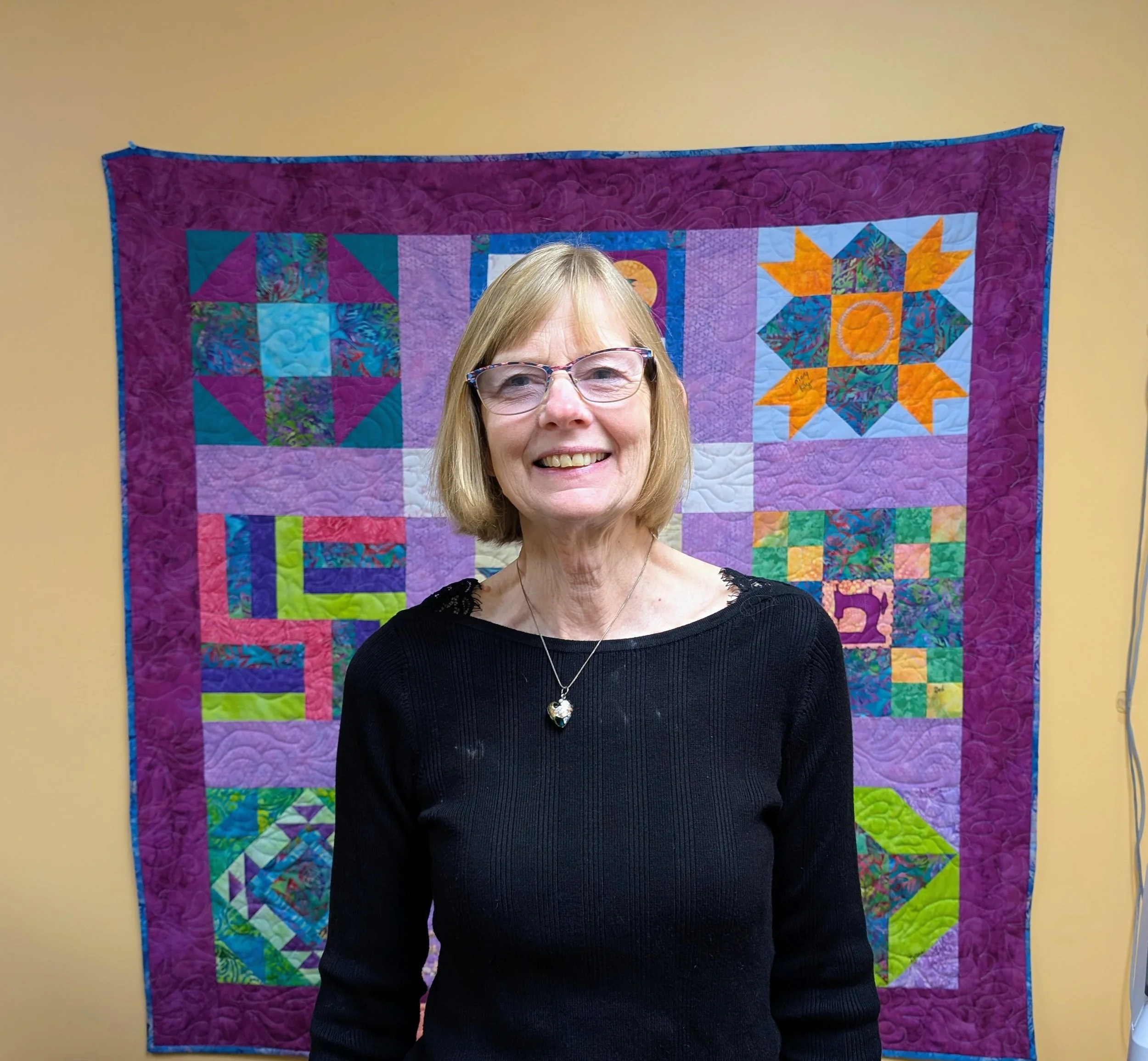 A smiling woman with blonde hair and glasses wearing a sleeveless floral shirt against a plain background.
