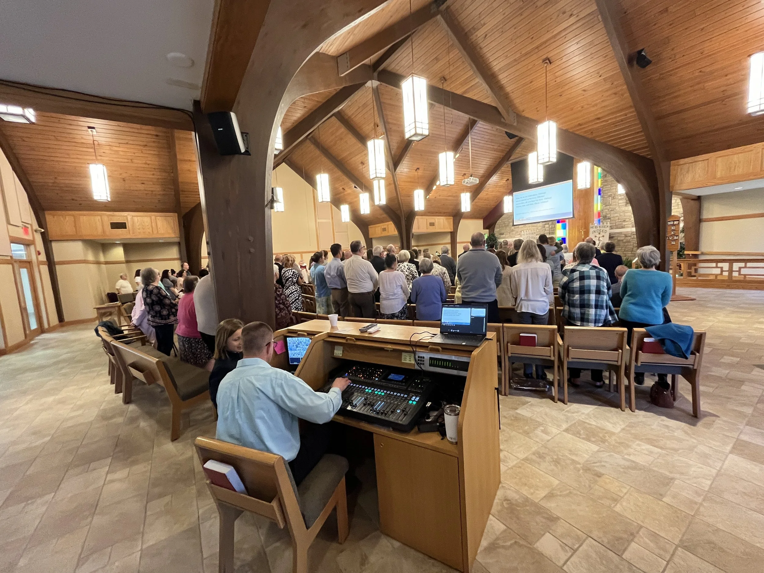 A large congregation of people standing in a church during service, with a worship leader or preacher at the front, a large screen displaying lyrics or scripture, and a person at the audio control panel in the foreground, all under a wooden cathedral ceiling with hanging lights.