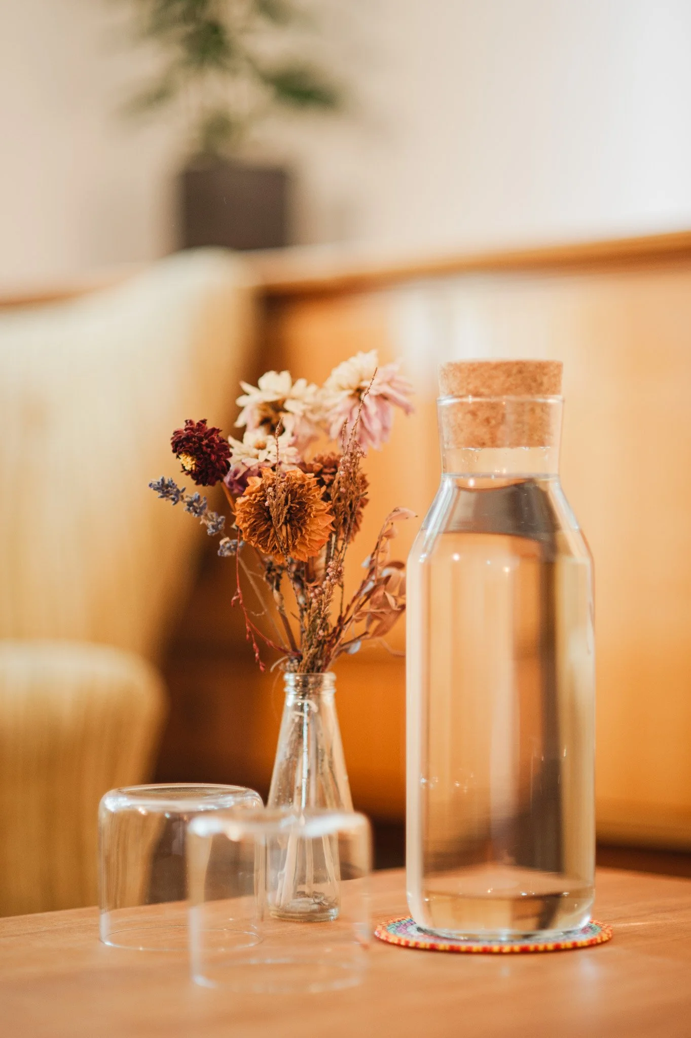 Vase mit getrockneten Blumen, Wasserkrug mit Korken, zwei leere Gläser auf Holztisch.