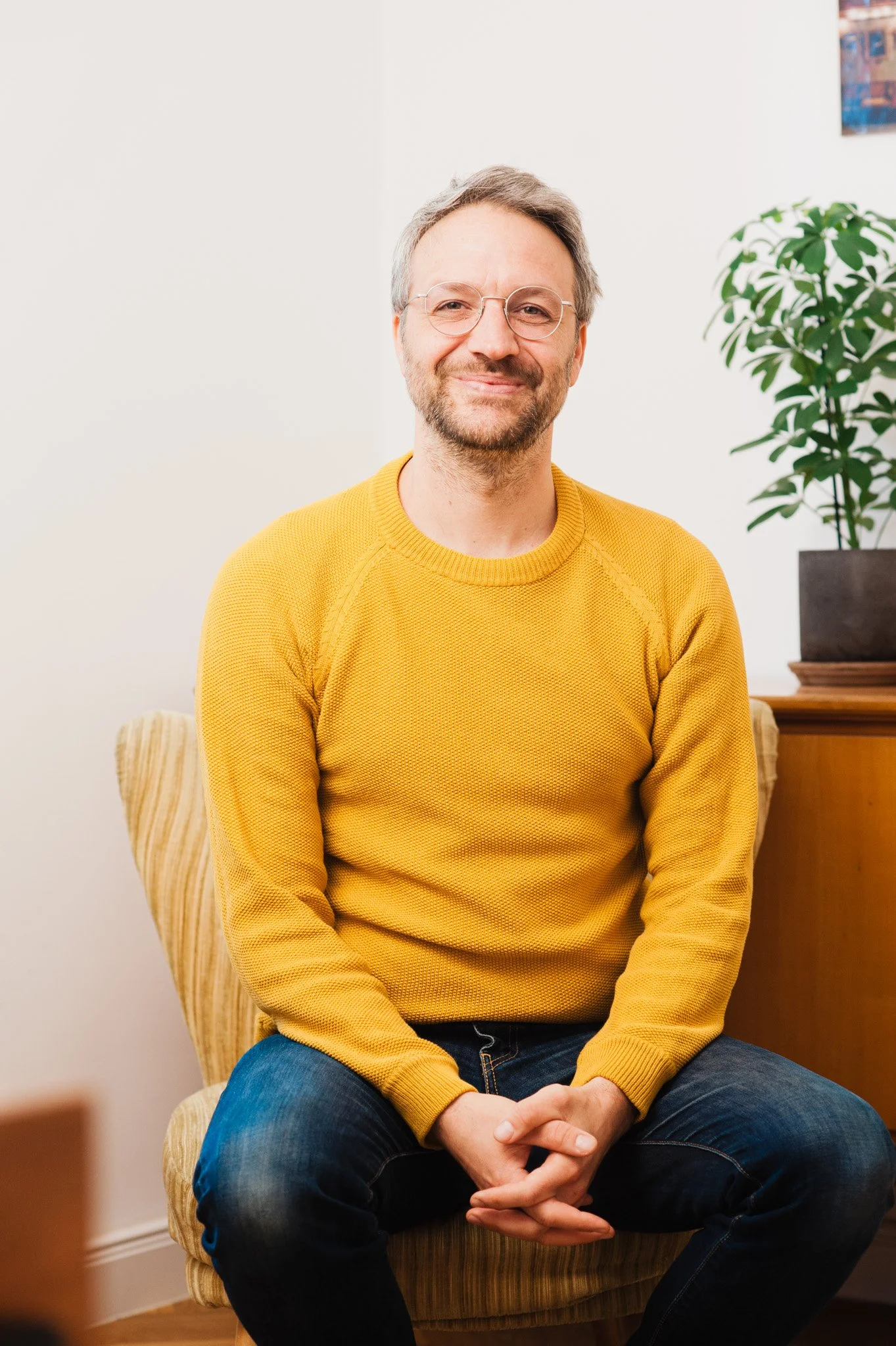 Ein Mann mit Brille, grauen Haaren und einem Bart sitzt auf einem gelben Sessel, trägt einen gelben Pullover und dunkelblaue Jeans, im Hintergrund eine Zimmerpflanze und eine weiße Wand.