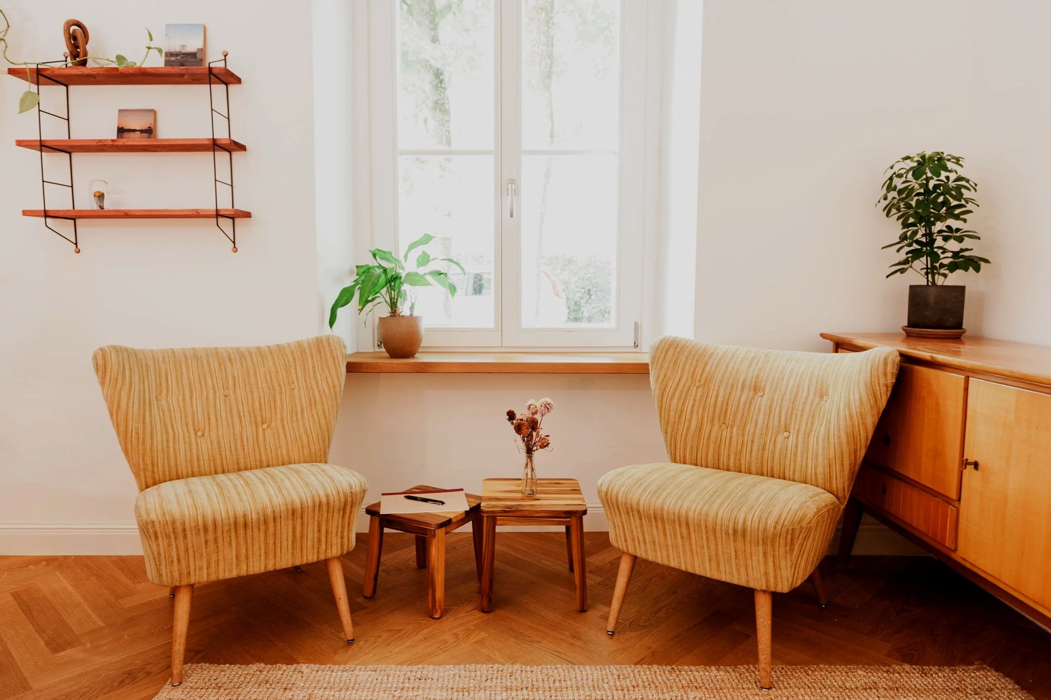Beratungspraxis in München: Gemütliches Beratungszimmer mit zwei Vintage-Sesseln, kleinen Beistelltischen, Pflanzen und großen Fenstern, die viel natürliches Licht einlassen.