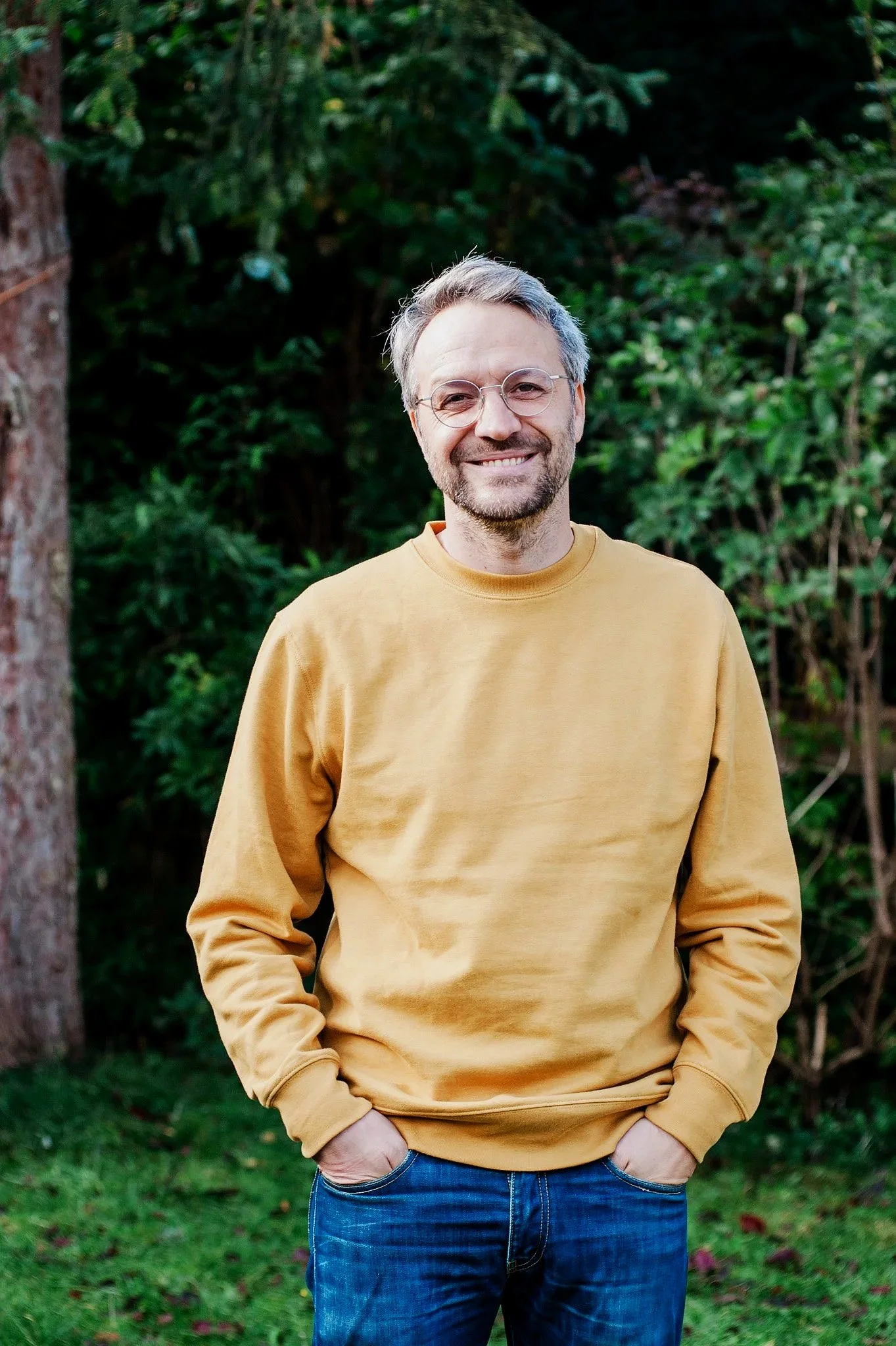 Ein Mann mit grauem Haar und Brille, trägt ein gelbes Sweatshirt und Jeans, steht vor grünen Büschen und Bäumen im Freien, lächelt in die Kamera.