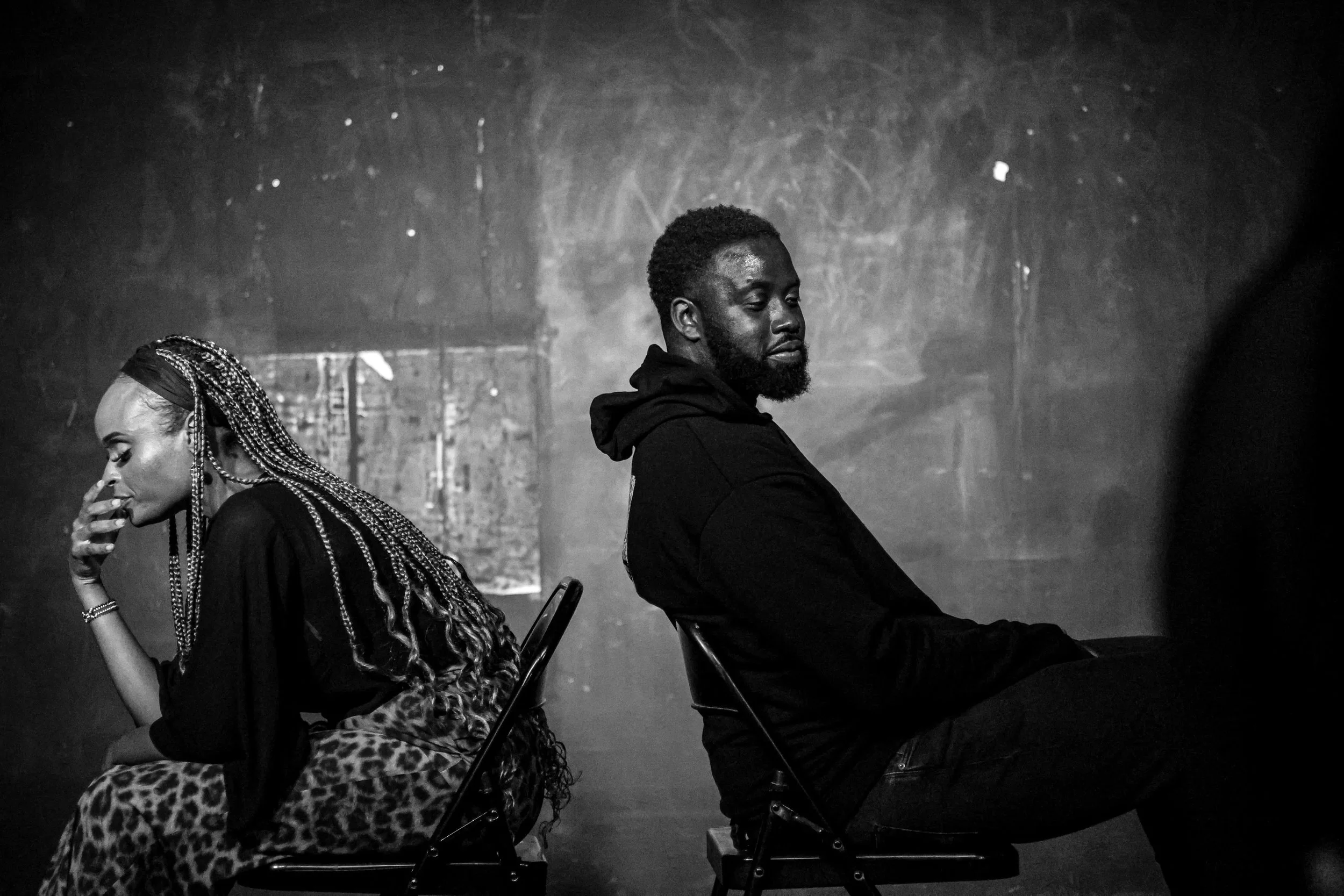 A black-and-white photo of a woman and a man sitting back-to-back on chairs, with their eyes closed. The woman has long, braided hair and is touching her face, while the man has a beard and is wearing a black hoodie.