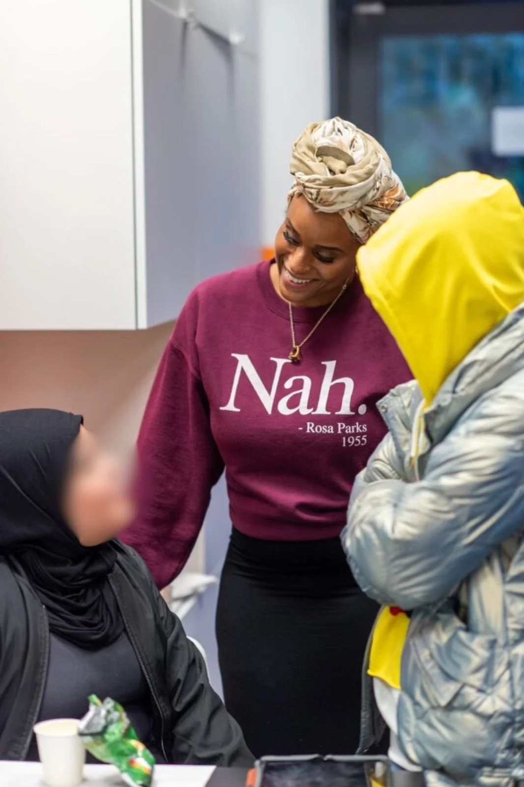 A woman wearing a maroon sweatshirt with 'Nah.' written on it, smiling and talking to two others in a casual indoor setting.