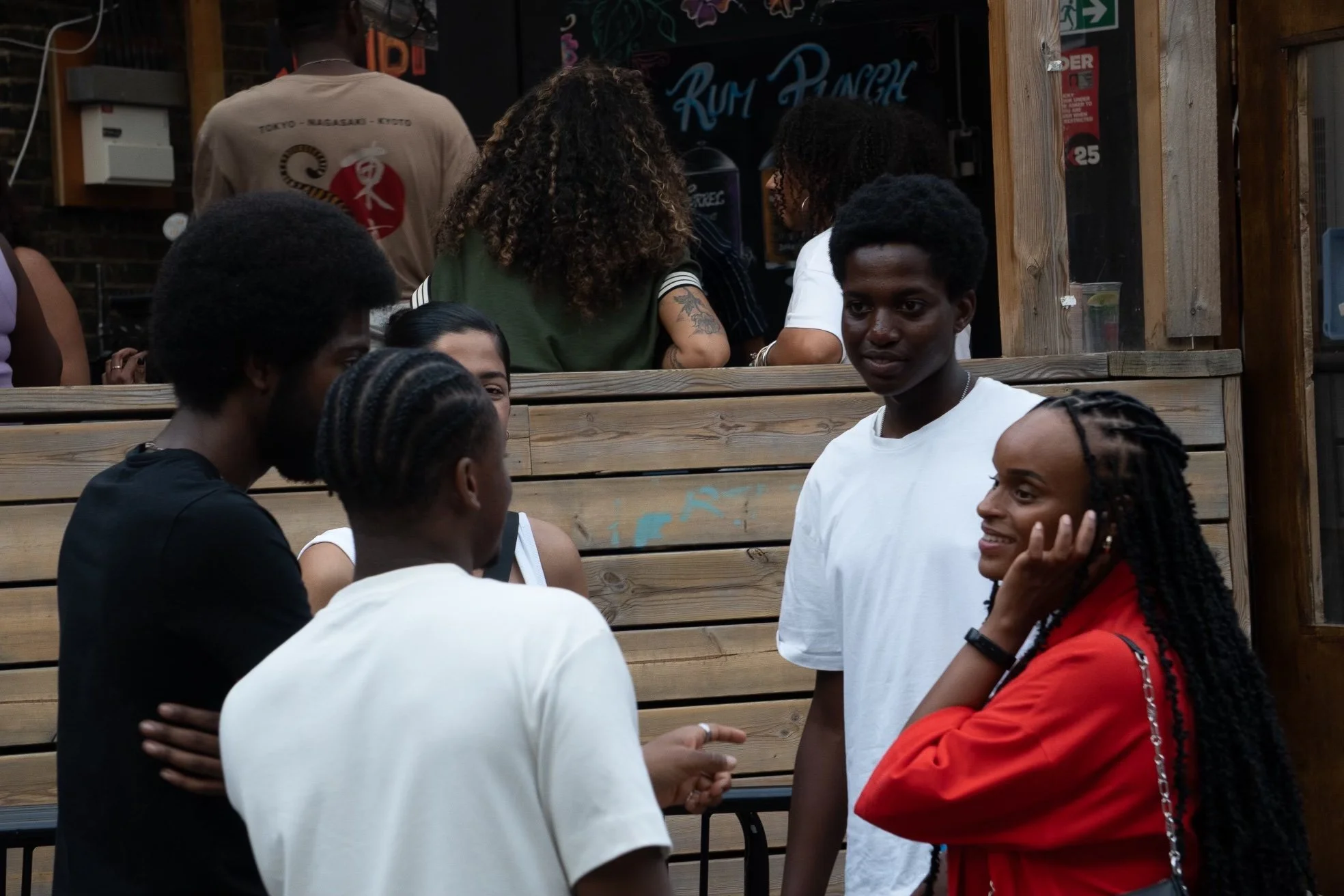 A group of five young people talking outdoors at a social gathering. They are standing in front of a wooden bar or fence, with some people in the background near a chalkboard sign and drinks.