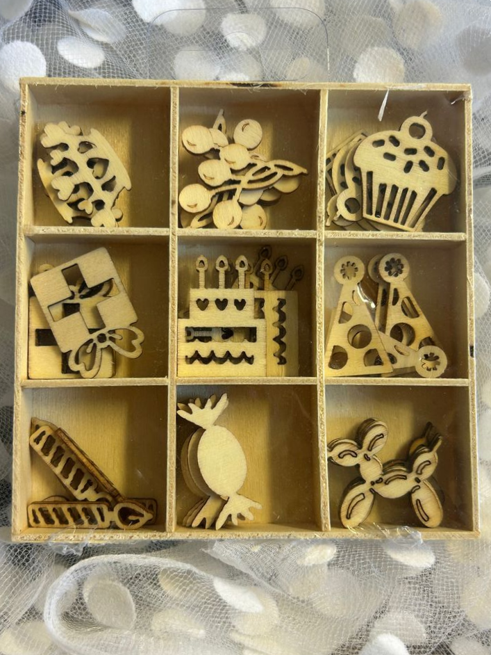 Boîte en bois avec neuf compartiments contenant des formes en bois de différentes décorations, comme des animaux, des gâteaux et des objets, sur un fond de tissu à motifs blancs.