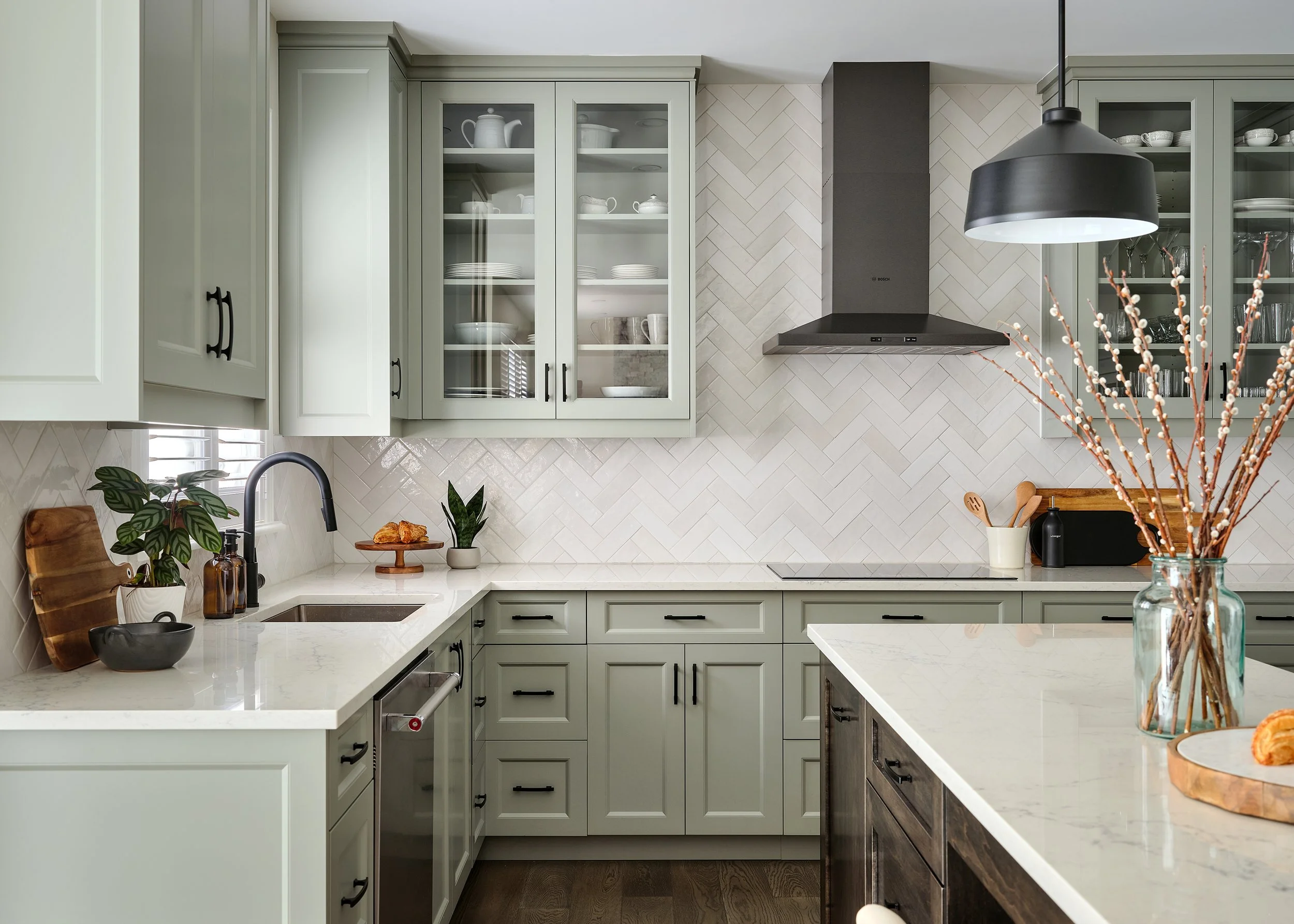 Custom kitchen renovation in Ottawa featuring warm green shaker cabinetry and glass cabinetry on either side of the exposed hood fan. 