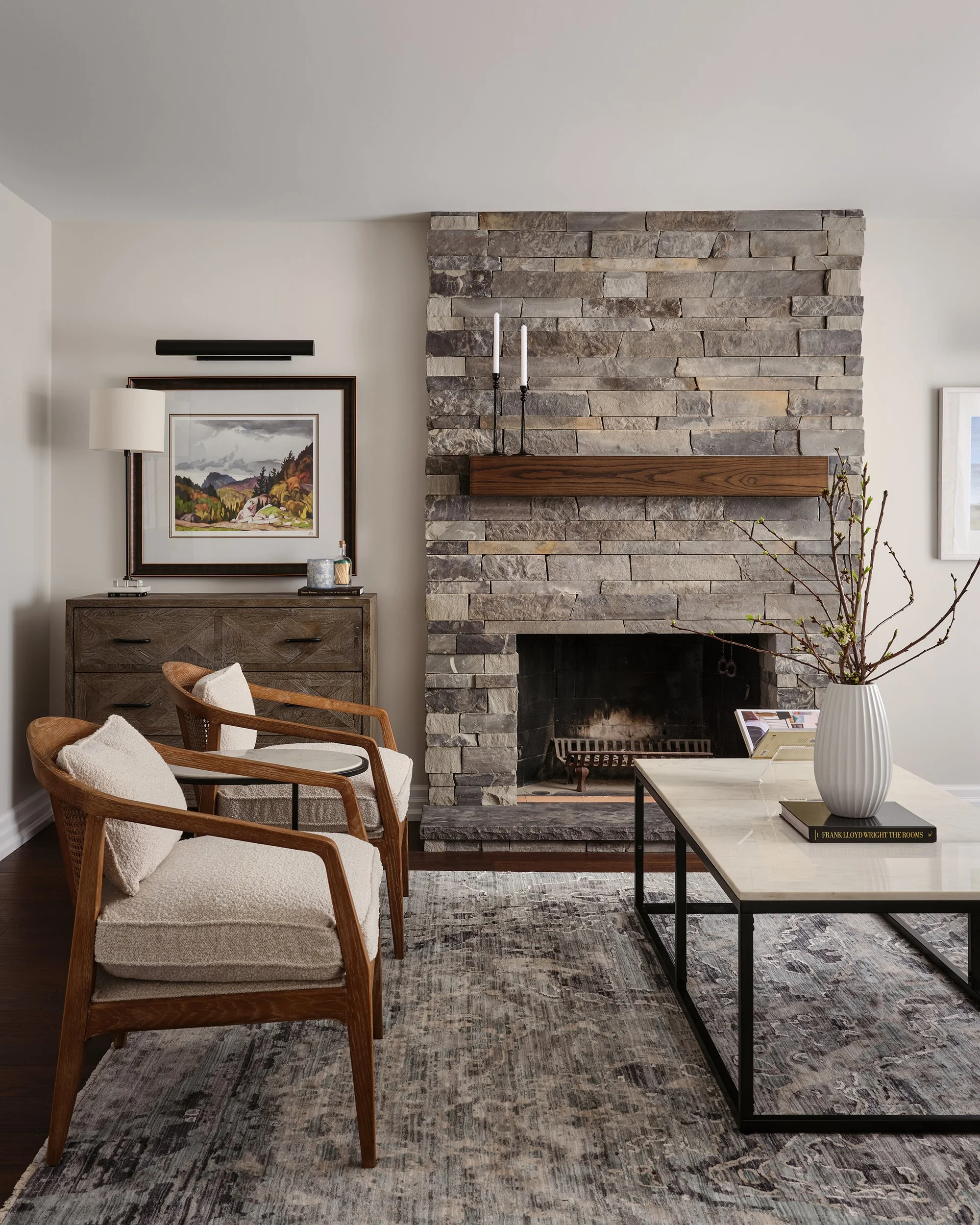 Living room with a stone fireplace, wooden mantel, two armchairs with cushions, a coffee table with a large vase of branches, a dresser with a lamp and framed art, and a patterned rug.