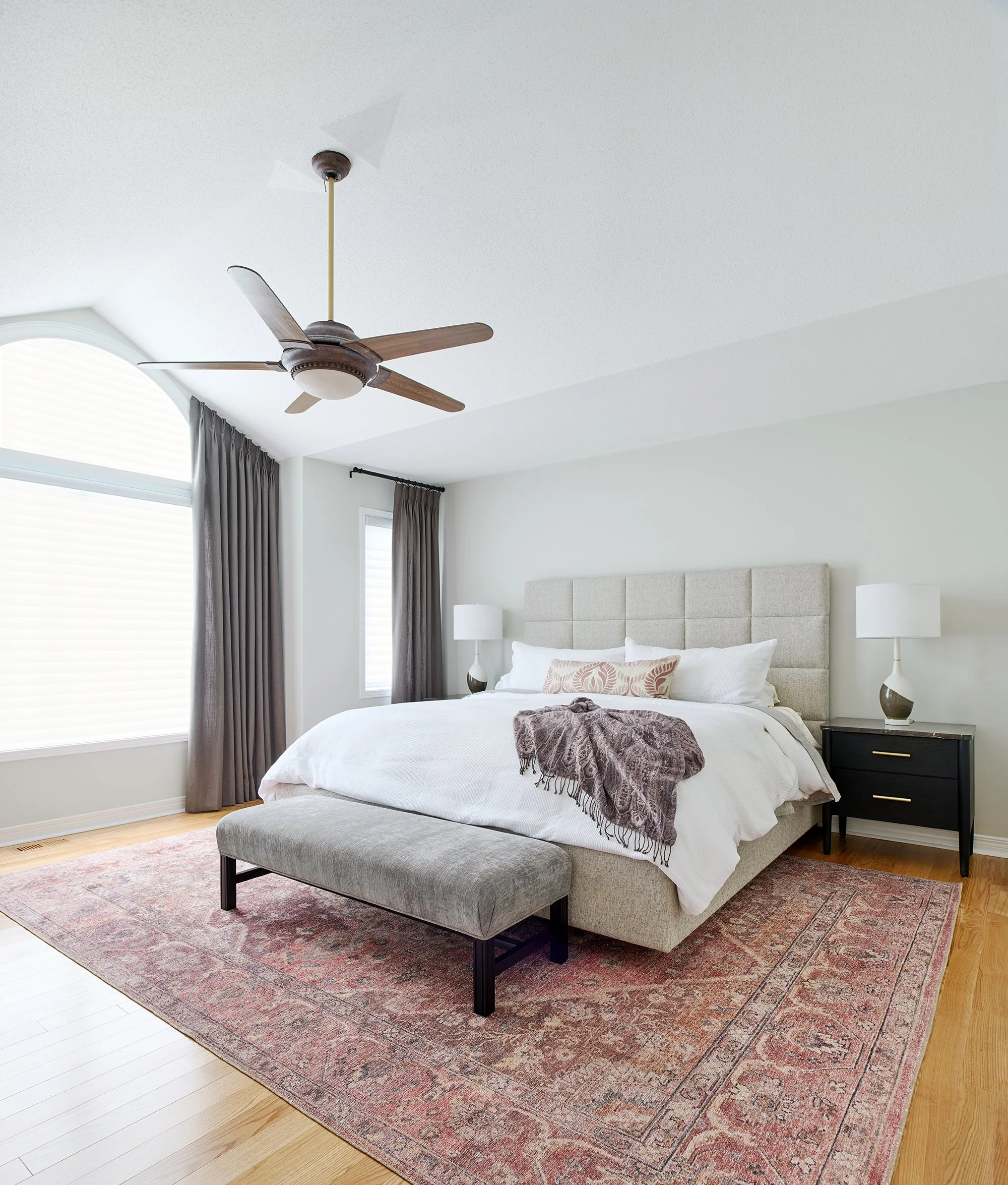 Principal bedroom design for an Ottawa family home featuring a pink area rug with a traditional pattern, upholstered bed frame and charcoal grey drapery. 