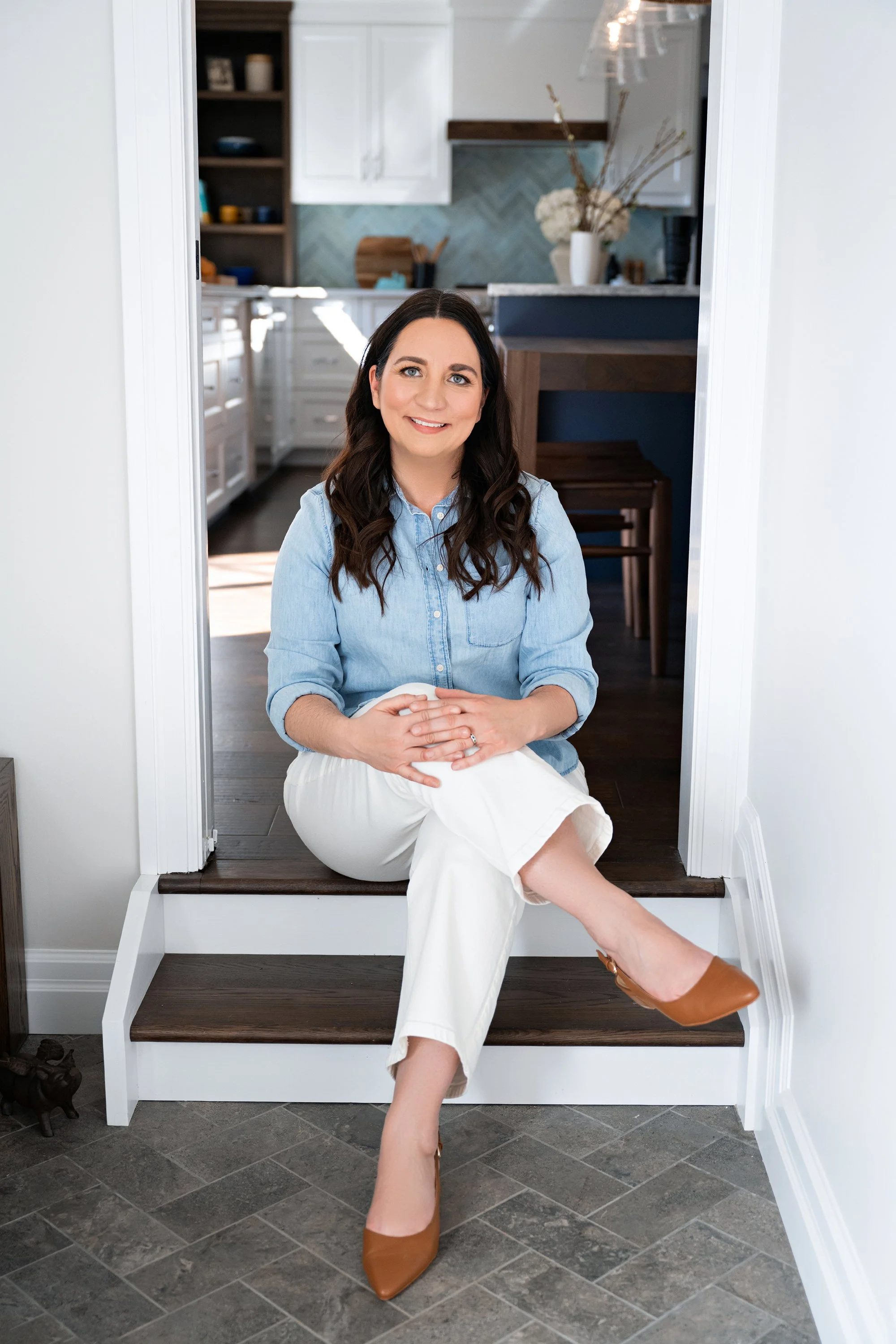 Natalie Cox, co-owner and Lead Designer of CPI Interiors, an Ottawa interior design firm is sitting on a step in front of a custom white kitchen