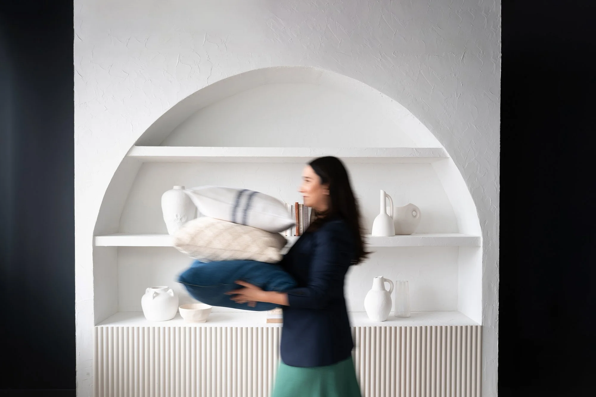 Natalie Cox, Co-owner and Lead Designer at CPI Interiors is carrying pillows in front of a white bookshelf.