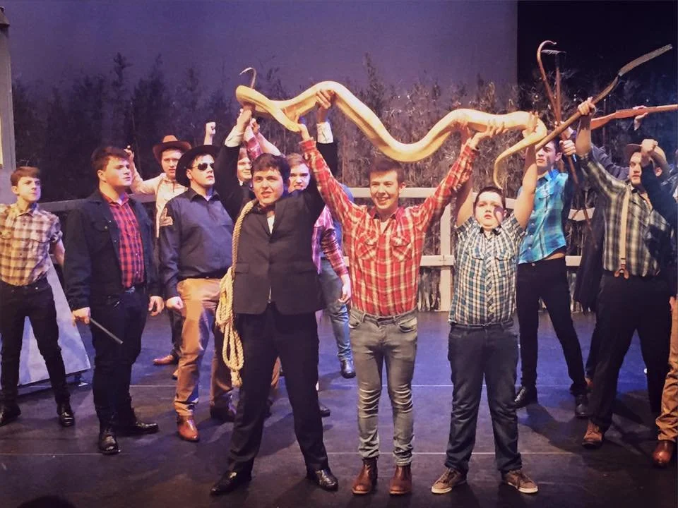 A group of young men on a stage wearing western-themed clothing, some holding sticks and a large snake, with a backdrop resembling a rural outdoor scene. Whistle Down the Wind