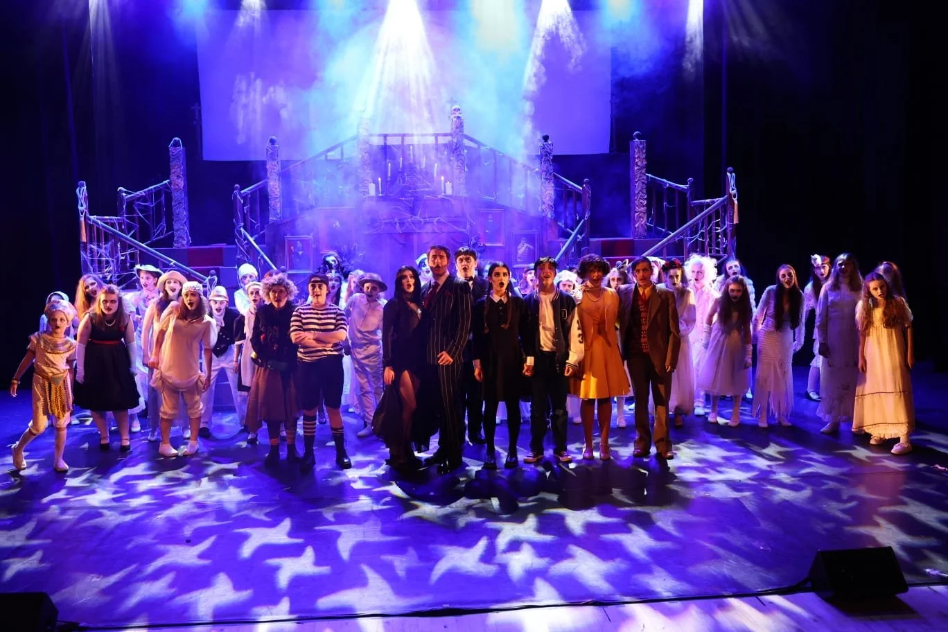 The cast of a theatrical performance on stage, dressed in period costumes, with a set resembling a grand staircase in the background, illuminated by colorful stage lights. The Addams Family