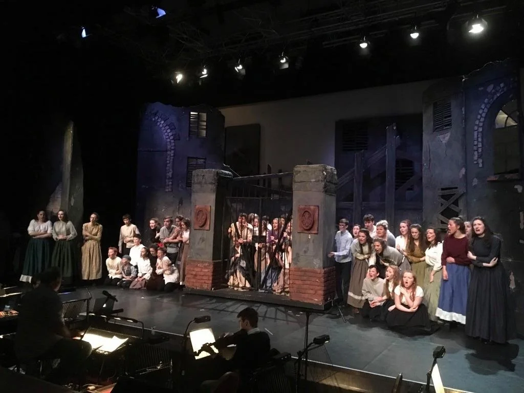 A large group of performers in period costumes on stage, with some singing and others in various poses, during a theatrical production of Les Mis
