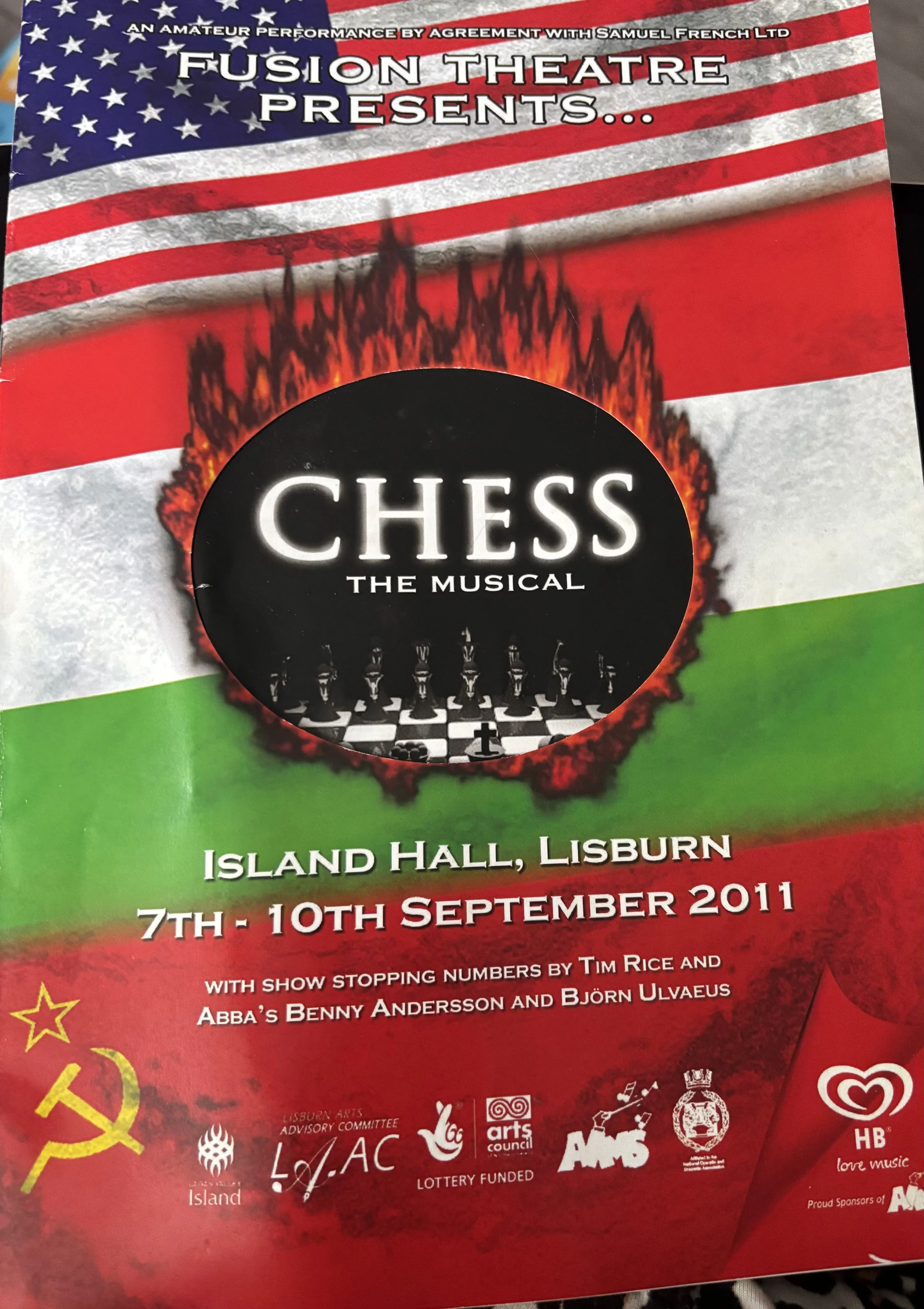 Black and white chessboard with three chess pieces, a red pawn, and checkered pattern with the text 'Chess: The Musical'.