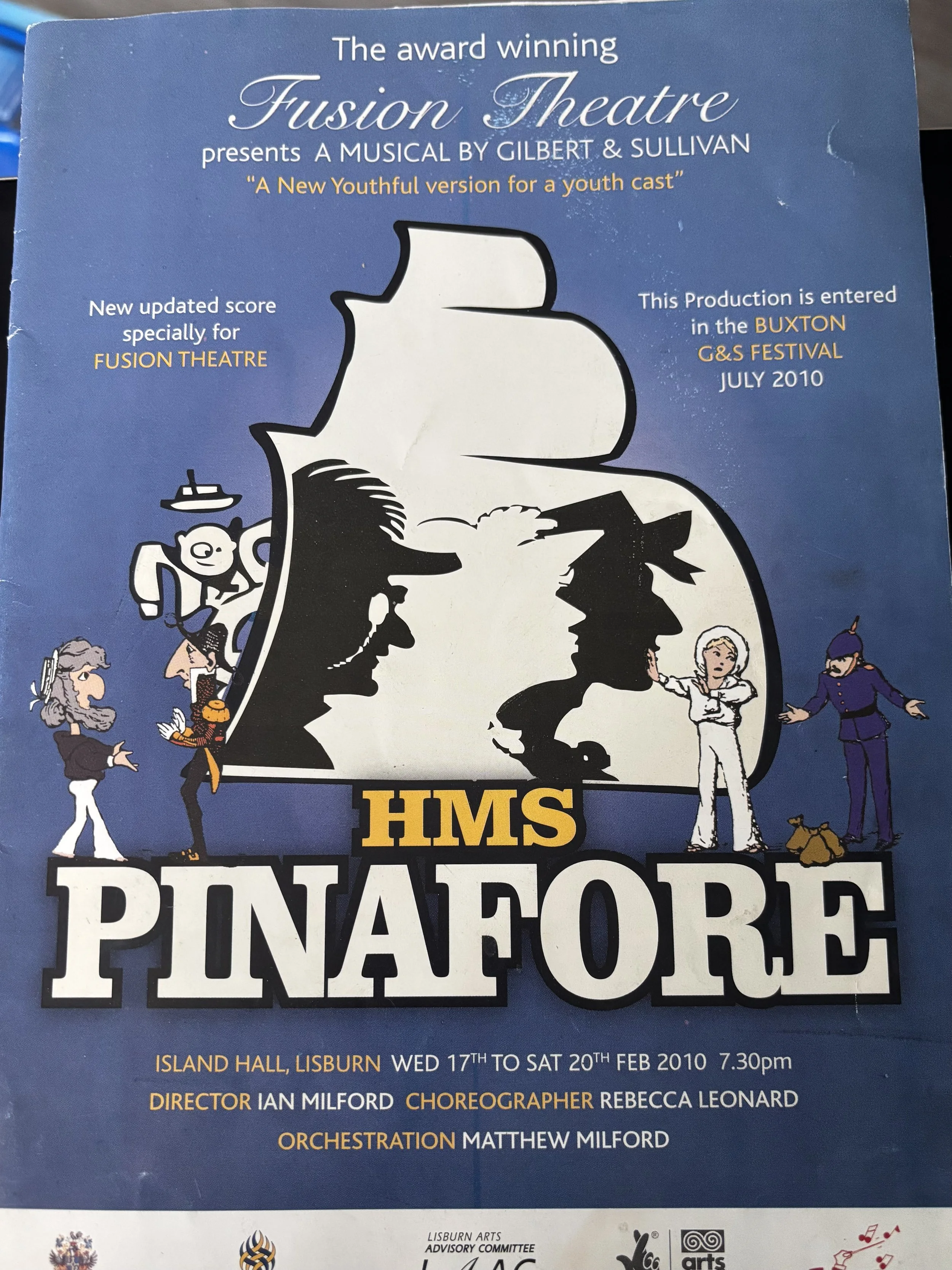 A festive poster featuring a cartoon sailor balancing a large wreath with the text 'HMS Pinafore' and 'Gilbert & Sullivan.'