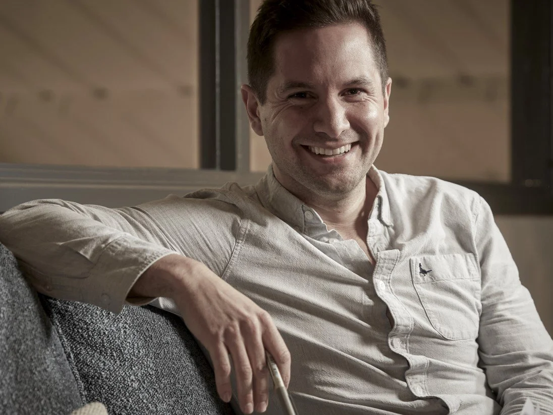 Smiling man sitting on a gray sofa, wearing a light-colored button-up shirt, with a casual and relaxed pose.