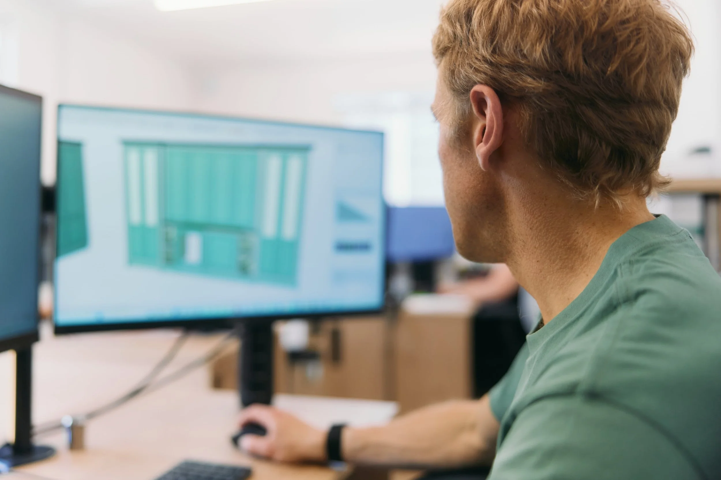A person with blond hair working at a computer in an office setting, viewed from behind.
