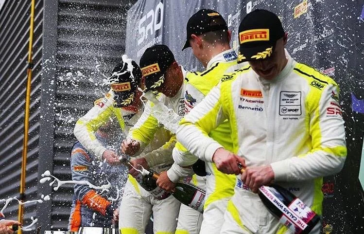 Race car drivers in racing suits and caps celebrating on the podium, spraying champagne, with a dark backdrop with sponsor logos.