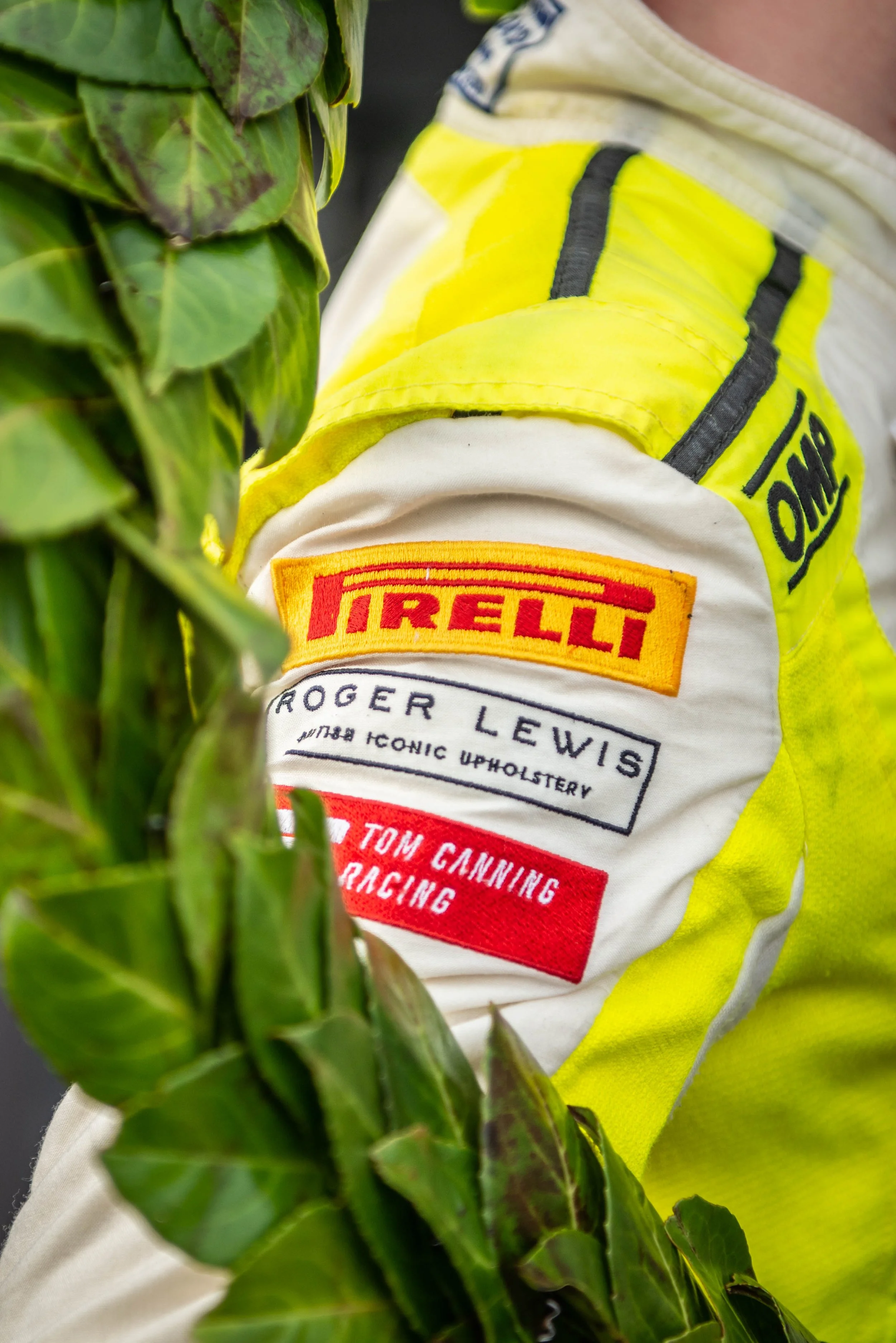 Close-up of a racing driver's arm showing sponsor patches on a white and yellow racing suit, with green leaves in the foreground.