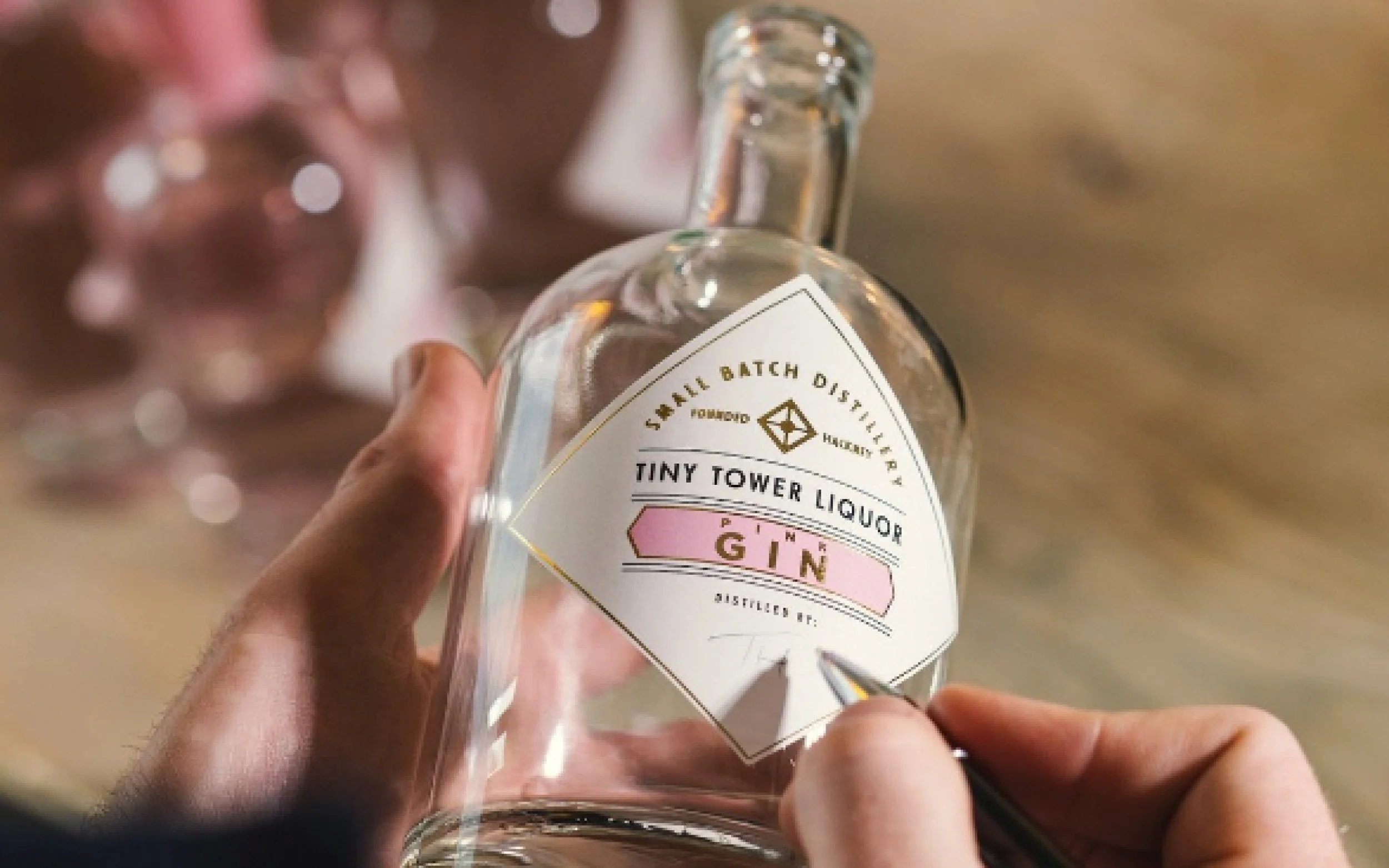A hand holding a clear glass bottle labeled 'Tiny Tower Liquor Gin,' with a person writing on the label.