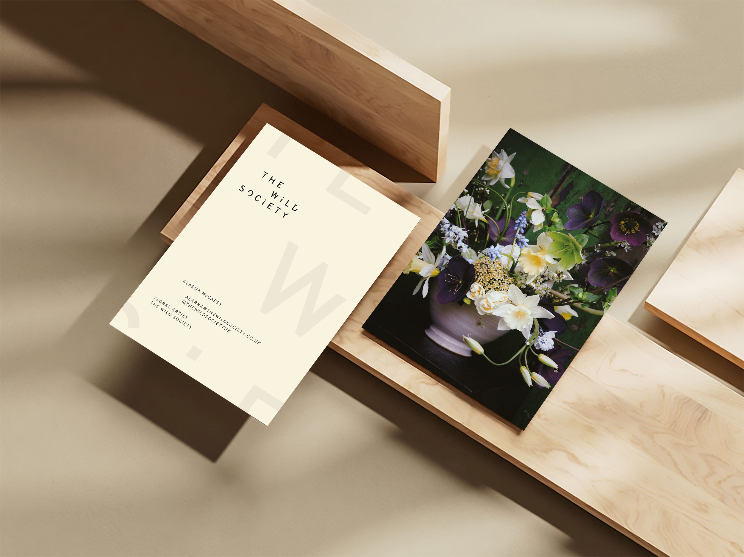A printed card with the words 'The Wild Society' and contact information next to a color photograph of a flower arrangement in a white bowl placed on a dark surface, all on a light-colored wooden table.