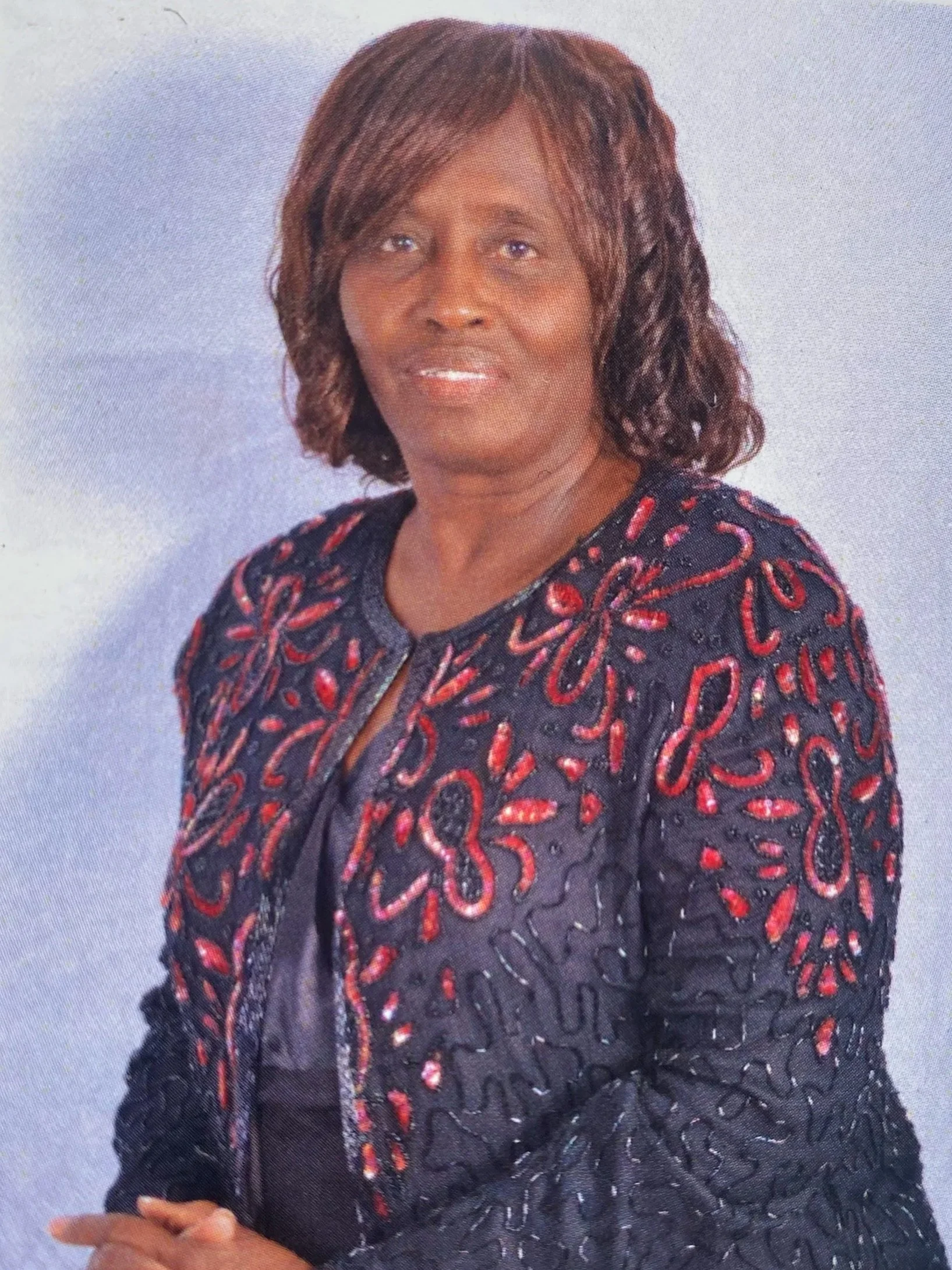 A woman with shoulder-length brown hair wearing a black jacket with red and silver embellishments, standing against a plain background.