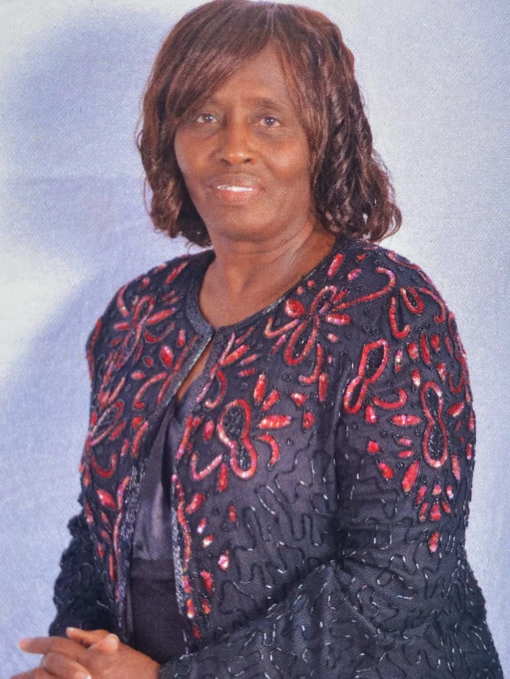A middle-aged African American woman with shoulder-length hair, wearing a black and red embroidered jacket, posing against a light-colored background.