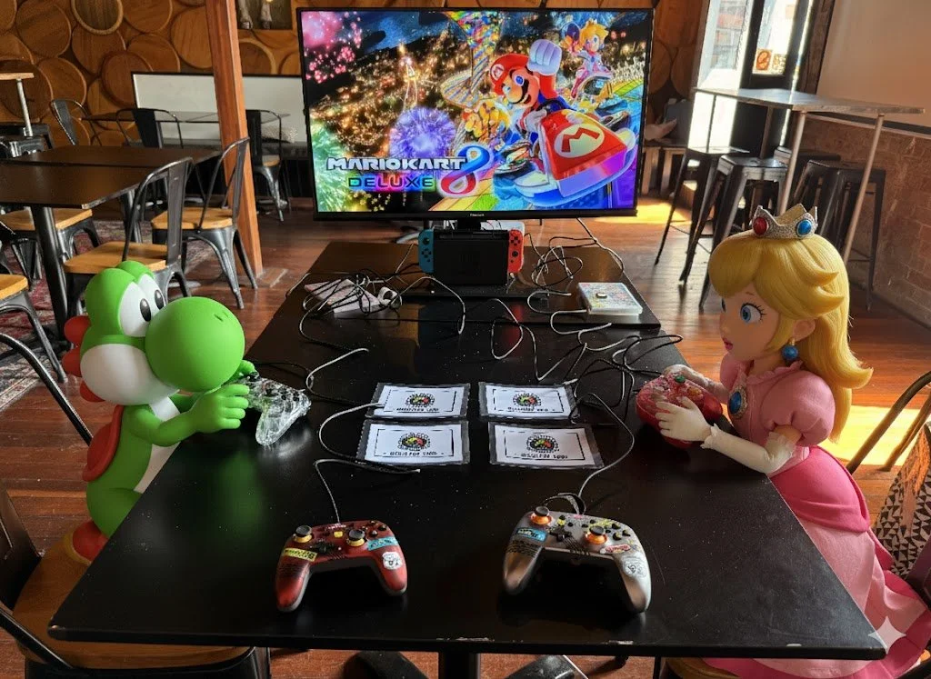 Yoshi and Princess Peach playing Nintendo Switch at a Mario Kart Deluxe 8 gaming setup with a large screen display of the game, in a cozy indoor setting.