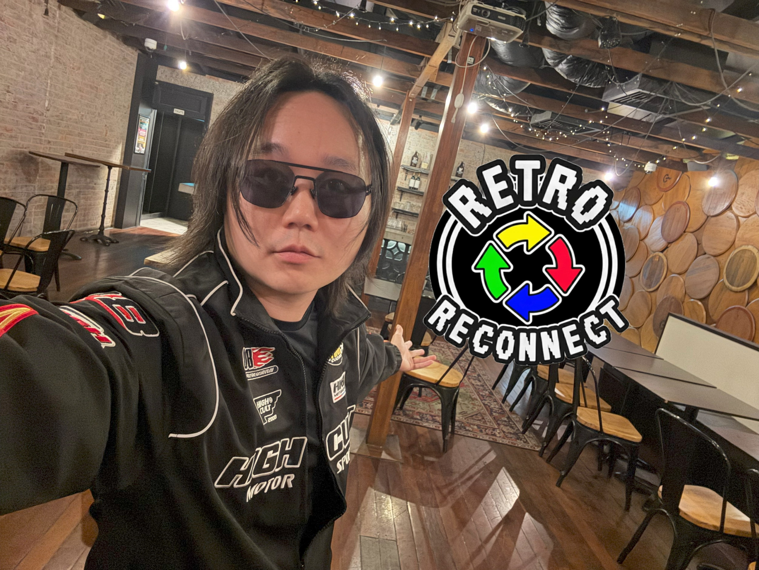 A person with shoulder-length black hair, wearing sunglasses and a racing jacket, takes a selfie in a decorated cafe or bar with a wooden interior. There is a large sign on the wall that reads 'RETRO RECONNECT' with colorful arrows and circular designs.