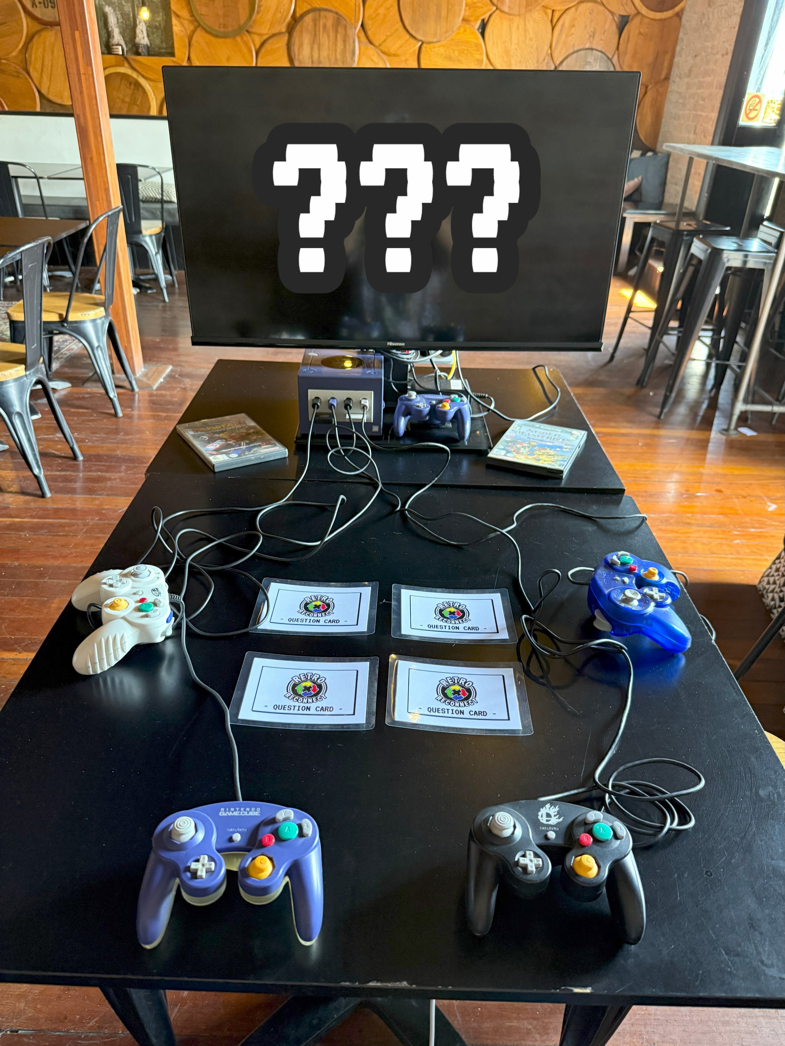 A gaming setup with a flat screen TV displaying three question marks, two game controllers (one purple and one blue), four printed question cards on the table, and a gaming console in a restaurant or café setting.