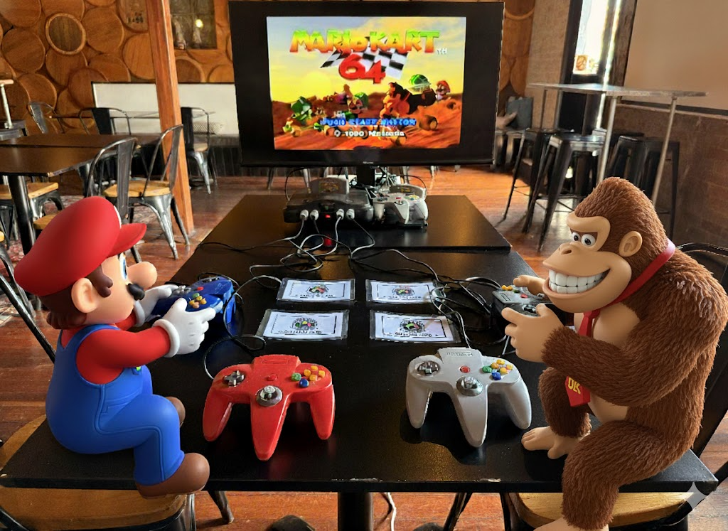 Mario and Donkey Kong plush toys playing Mario Kart 64 video game with controllers at a table, with a TV screen displaying the game's title in the background.