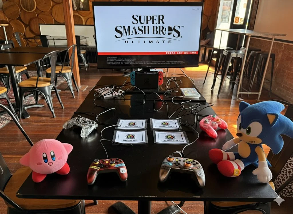 A gaming setup on a black table with a large TV displaying the Super Smash Bros. Ultimate logo. The table has four game controllers, including two Sonic the Hedgehog plush toys, a pink Kirby plush, and some identification cards or papers. The backgro