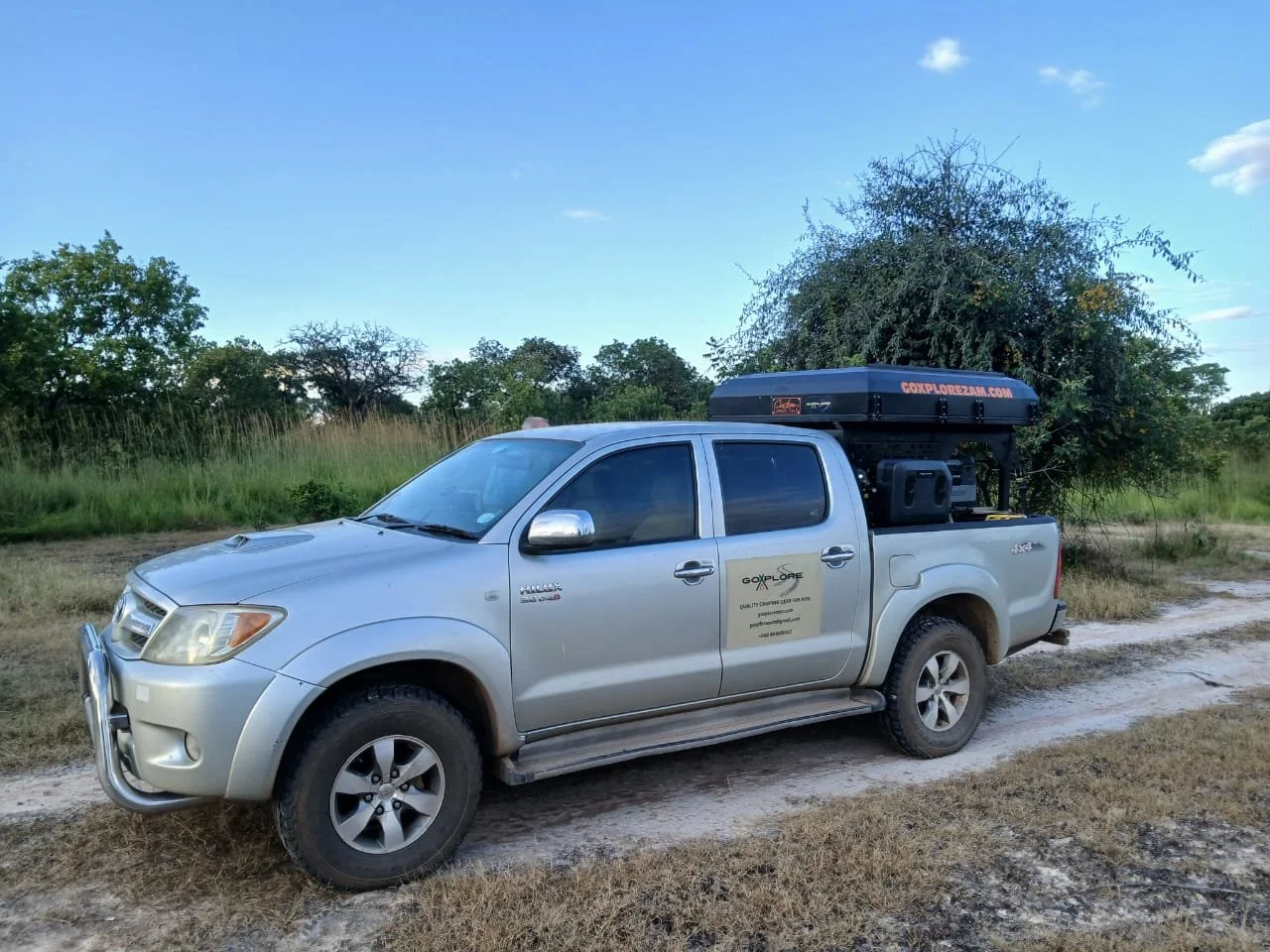 A silver pickup truck parked on a dirt road in a natural environment with green trees and grass, with a black rooftop cargo box and a company logo on the door.