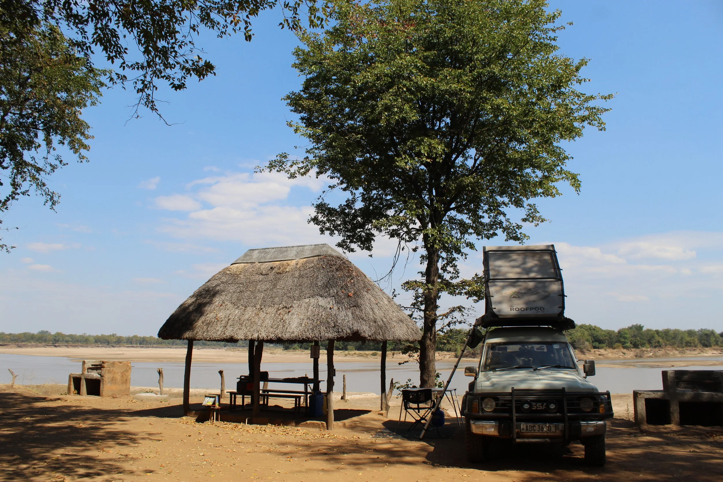 A thatched roof hut with a tree next to it, a vehicle with a rooftop tent labeled 'ROOFPOD,' a small seating area under the tree, and a water body in the background under a partly cloudy blue sky.