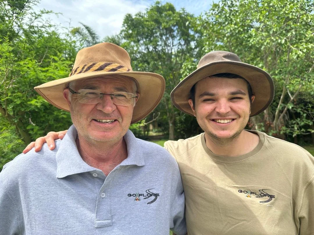 Two men outdoors, smiling, wearing wide-brimmed hats and casual shirts with a logo, surrounded by green trees.