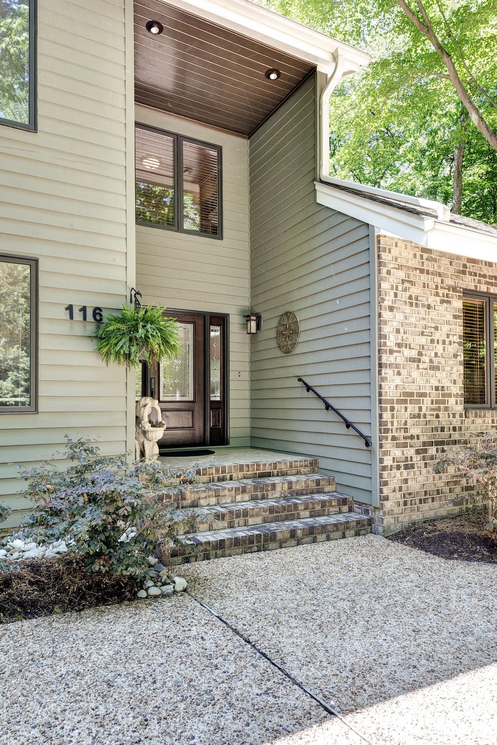 The front entrance of a house with a dark brown door, set on brick steps, with house numbers 116 on the beige siding, surrounded by greenery and a hanging plant to the left of the door.
